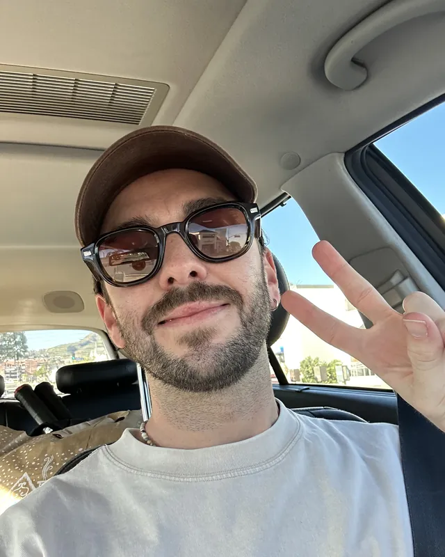 Tyler in a car, in a cap and sunglasses, making a peace sign