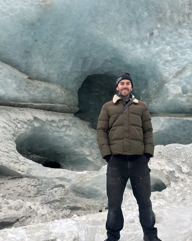 Tyler standing in front of an ice cave in a glacier, in a beanie and a puffer jacket